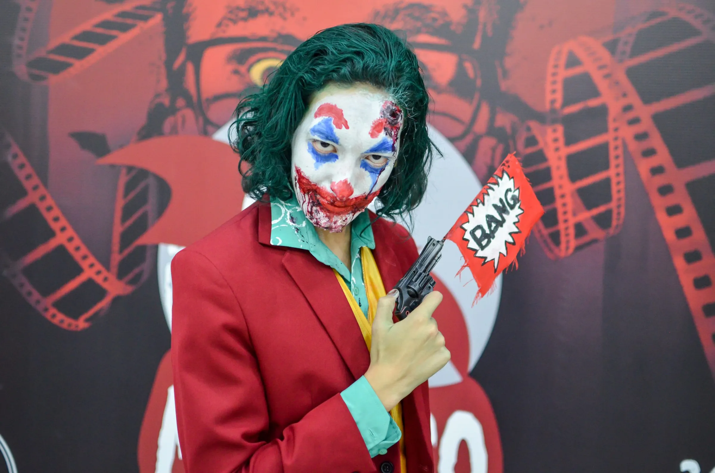 Cosplay de Coringa com o rosto pintado e uma arma de brinquedo com a bandeira BANG.